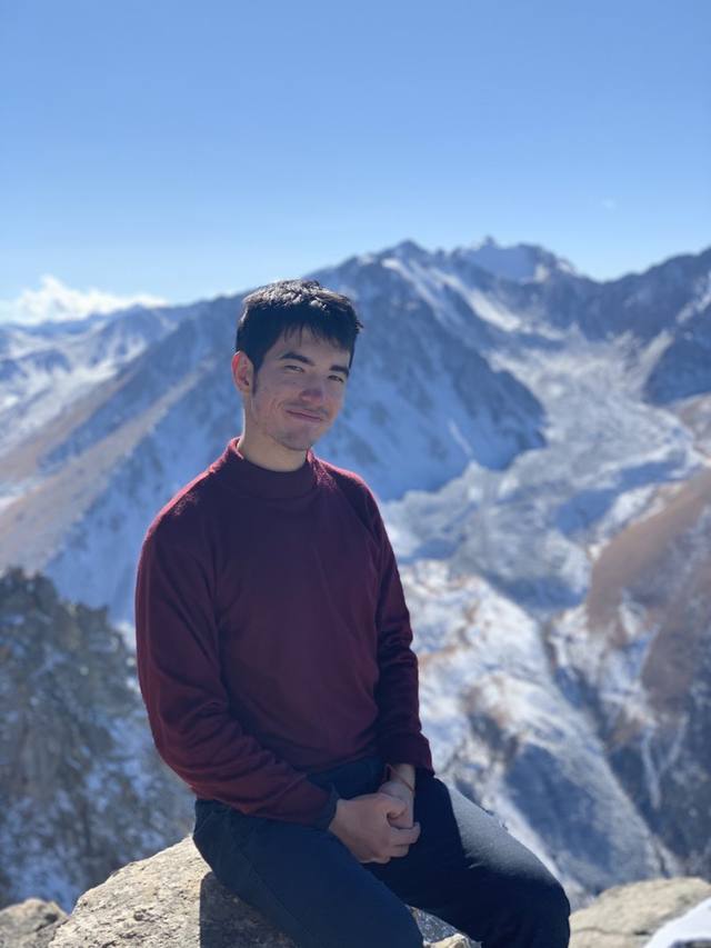 Alisher sitting on a rock in the mountains, snowy peaks behind him