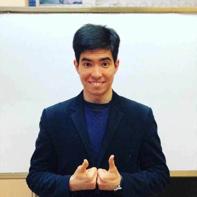 Alisher smiling with two thumbs up in front of a whiteboard